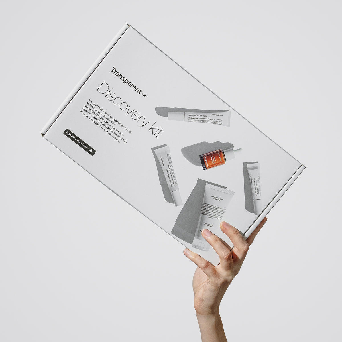 Hand hält eine „Discovery Kit Transparent Lab“-Box mit Hautpflegeprodukten vor einem schlichten Hintergrund.