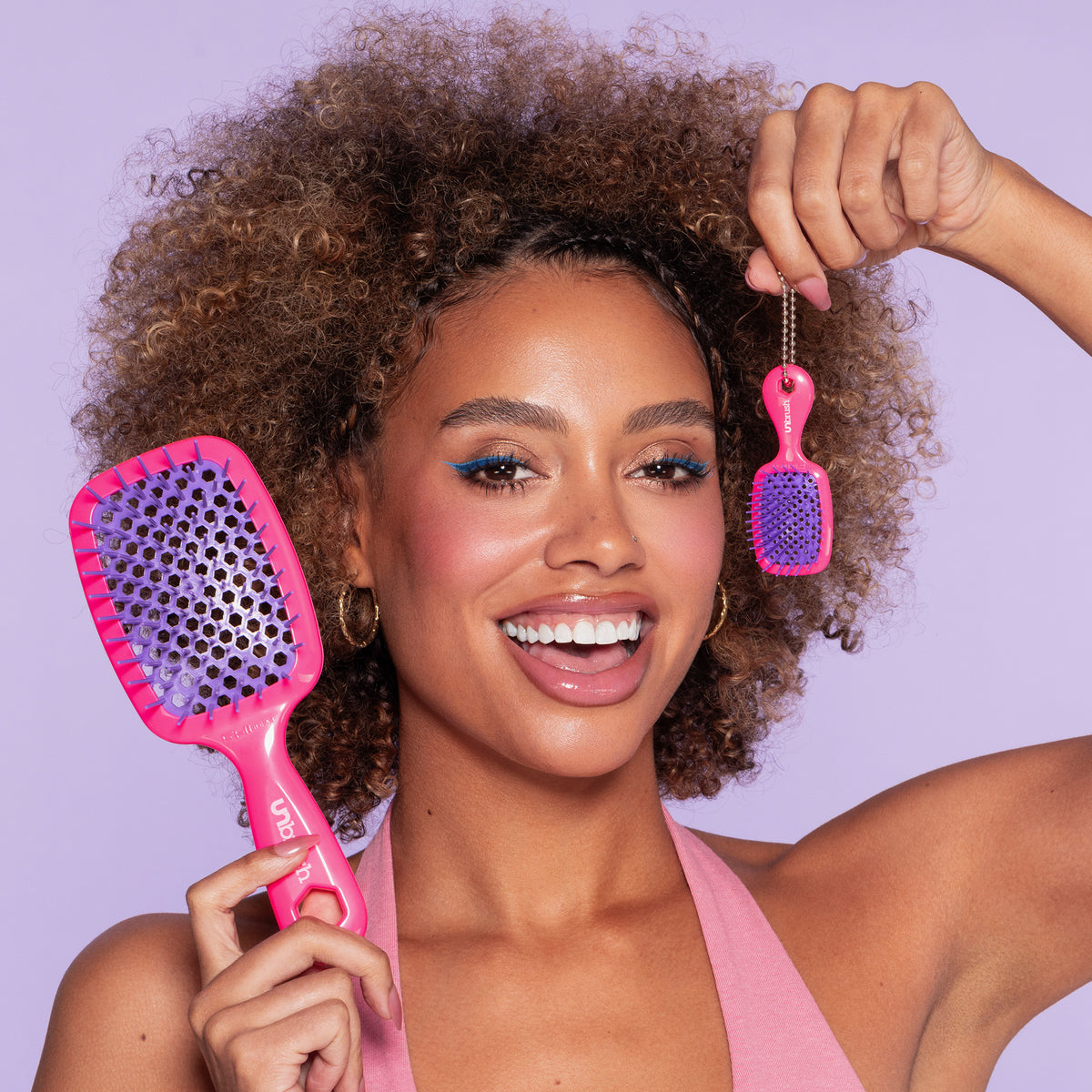 Frau mit lockigem Haar lächelt und hält eine große pink-lila UNbrush® Bürste sowie eine Mini-Version am Schlüsselanhänger hoch.