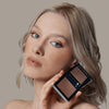 Eine Frau hält den geöffneten Skin Mate Compact von Mesauda Beauty in der Nähe ihres Gesichts und blickt in Kamera.