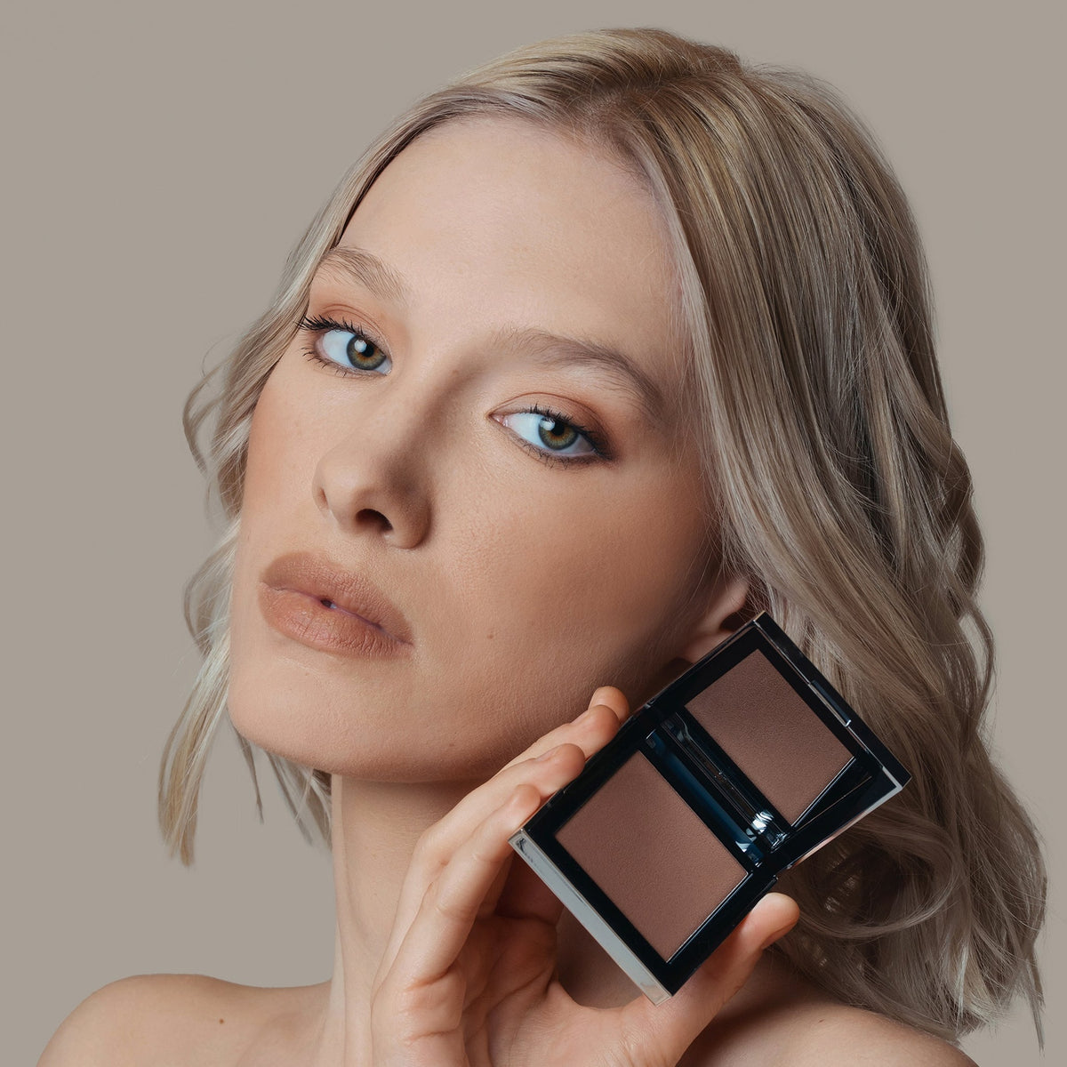 Eine Frau hält den geöffneten Skin Mate Compact von Mesauda Beauty in der Nähe ihres Gesichts und blickt in Kamera.