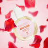 Runde Flasche Hair Syrup Rose Bloom Haaröl schwebt vor rosa Hintergrund, umgeben von roten Rosenblättern; Etikett und Text klar sichtbar.