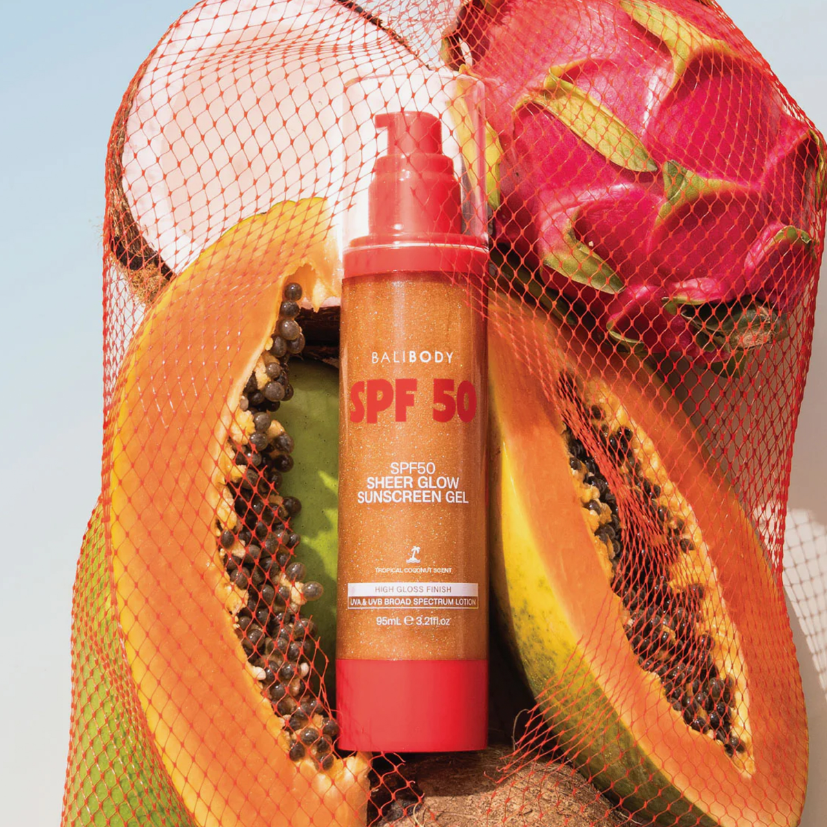 Ein „Bali Body Sheer Glow Sunscreen Gel“ LSF 50 mit Papaya-Hälften und Drachenfrucht unter einem roten Netz.