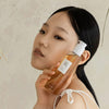 Eine Person mit langem dunklem Haar und neutralem Gesichtsausdruck hält eine Flasche Ginseng Cleansing Oil an ihre Wange