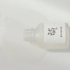 Eine liegende Flasche Glow Deep Serum: Rice + Alpha Arbutin mit heraustretender transparenter Flüssigkeit auf hellem Hintergrund