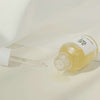 Eine liegende Flasche Glow Serum: Propolis+Niacinamide, aus der die hellgelbe Flüssigkeit auf eine weiße Oberfläche tritt