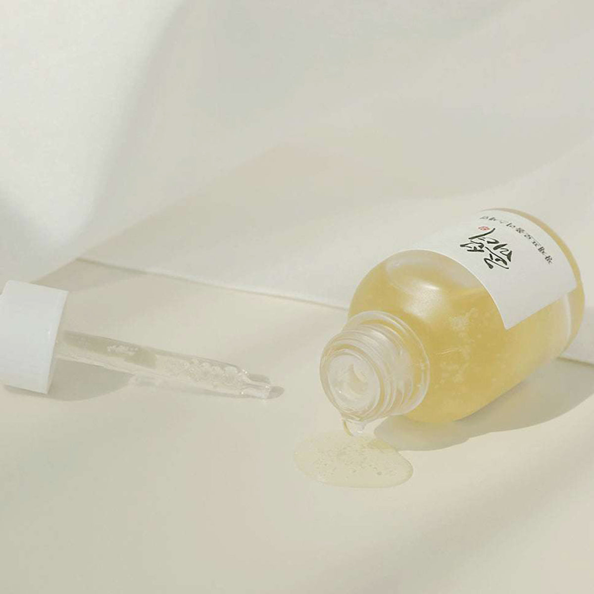 Eine liegende Flasche Glow Serum: Propolis+Niacinamide, aus der die hellgelbe Flüssigkeit auf eine weiße Oberfläche tritt