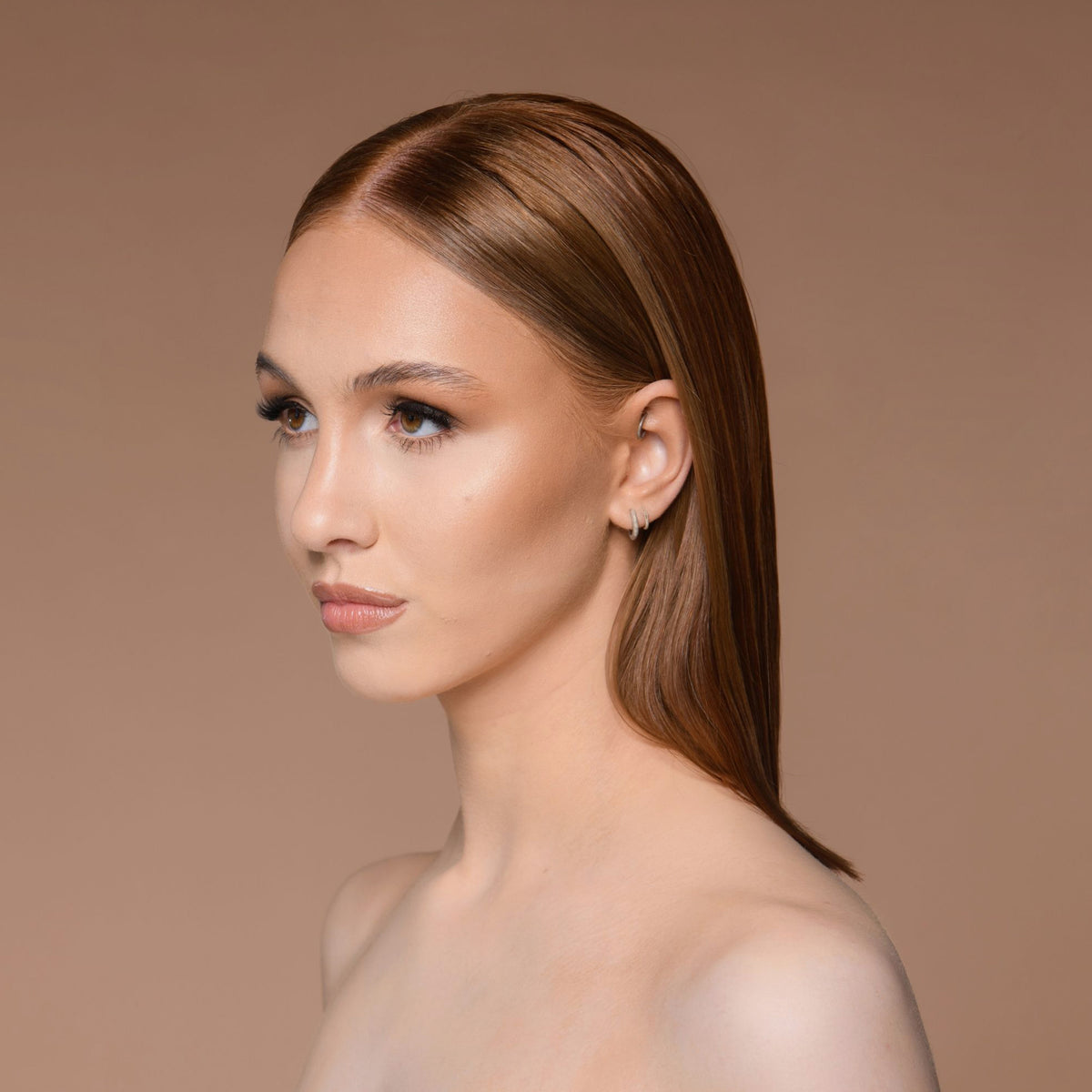 Eine Frau mit glattem braunem Haar und dezentem Make-up, aufgepeppt mit Cronzer – Cream Bronzer, blickt vor einem beigen Hintergrund zur Seite.