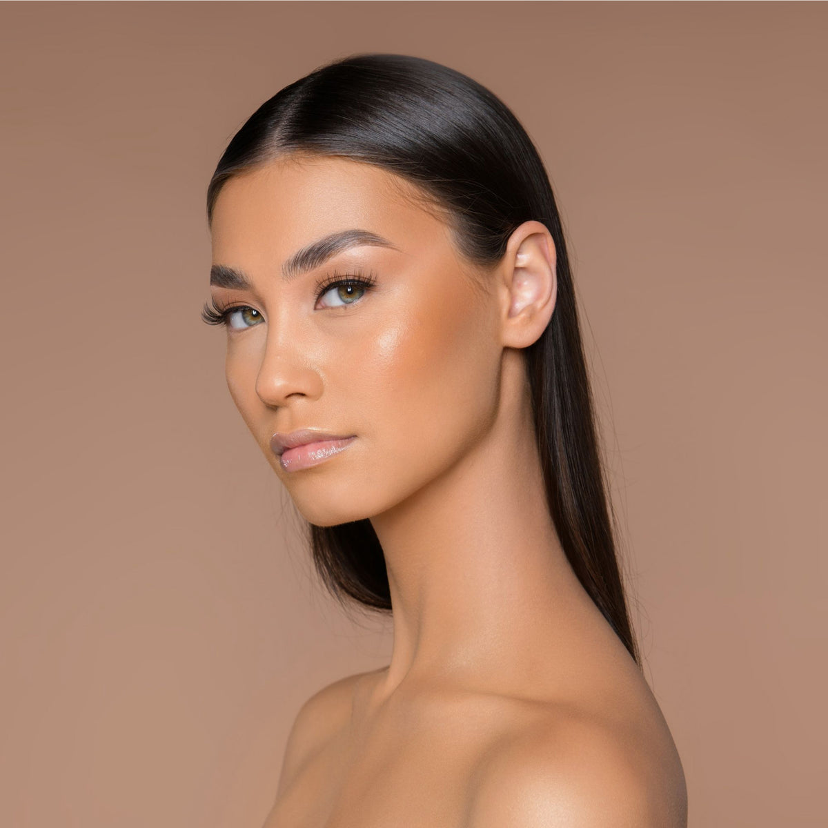 Eine Frau mit glattem dunklem Haar und strahlender Haut, mit freundlicher Genehmigung von Cronzer – Cream Bronzer, blickt vor einem sanften braunen Hintergrund über ihre Schulter.