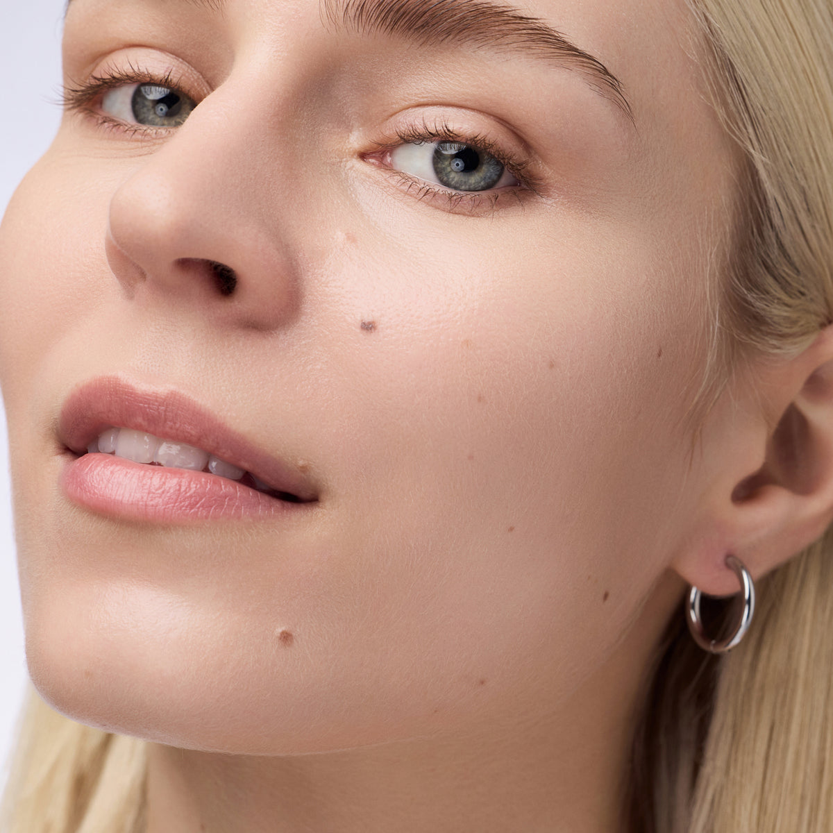 Nahaufnahme einer Person mit blondem Haar und hellblauen Augen die den Instant Bright Serum Concealer 001N auf der Haut präsentiert.