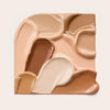 Mehrere ineinander fließende Swatches des Soft Glam Filter Fluid in Braun-, Beige- und Cremetönen auf cremefarbenem Hintergrund.
