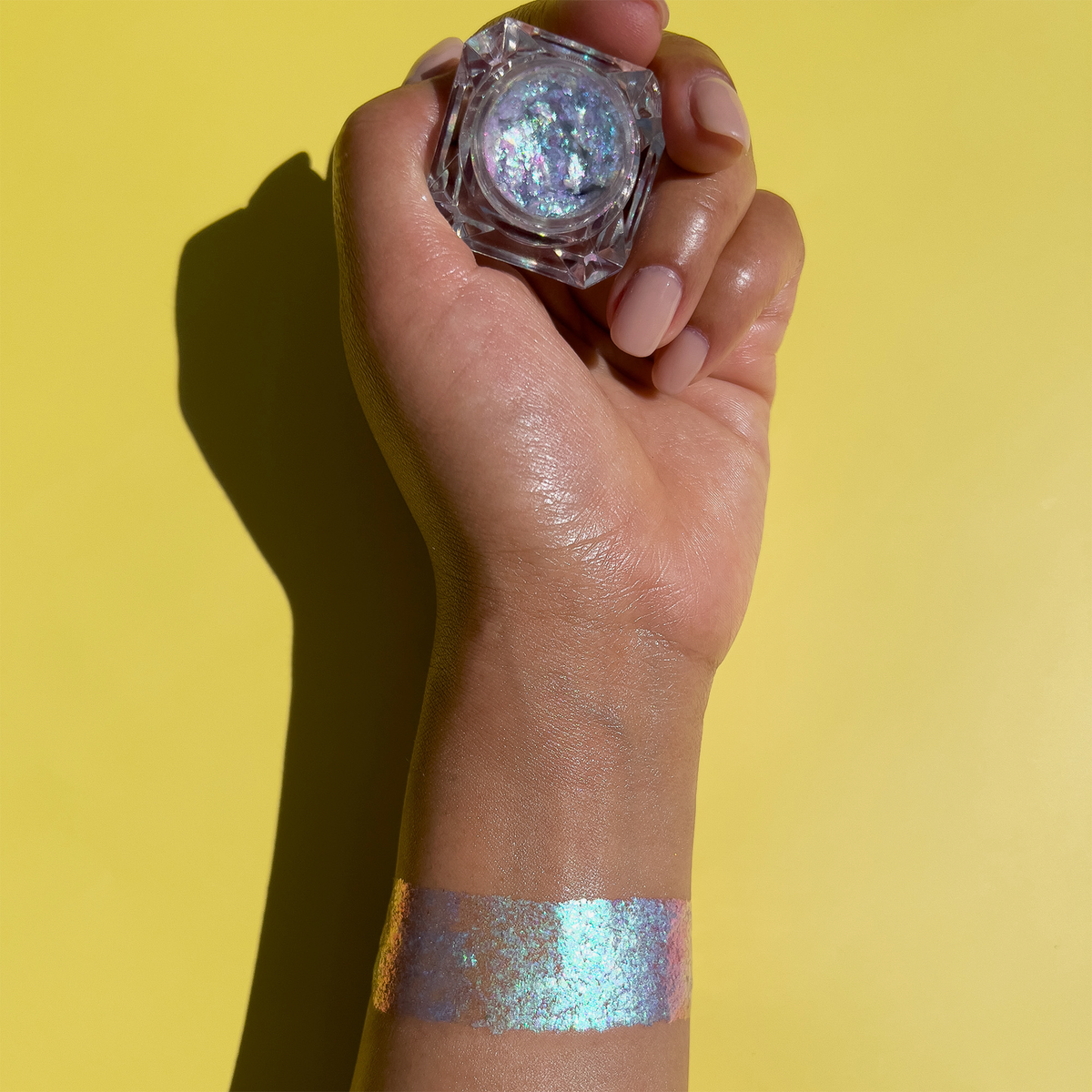 Eine Hand hält eine kleine Dose Danessa Myricks Beauty Infinite Chrome Flakes Pixie Dust mit einem Swatch am Handgelenk vor einem gelben Hintergrund.