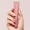 Eine Hand mit rosa manikürten Nägeln hält einen Dominique Cosmetics Juicy Lip Balm vor einem hellen Hintergrund.