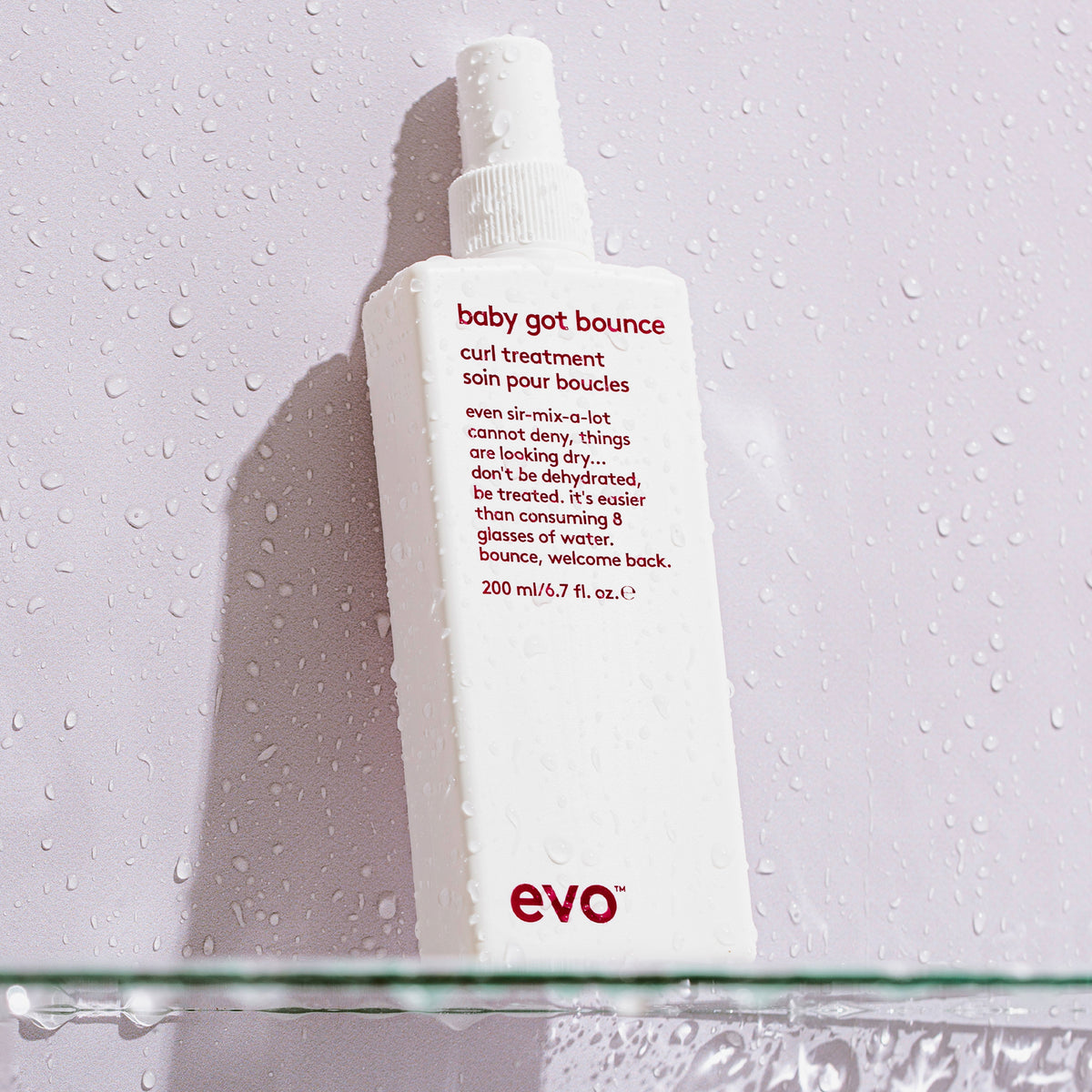 Die Flasche Baby Got Bounce Curl Treatment, weiß mit rotem Text, steht auf einer nassen Glasfläche mit Wassertropfen.
