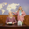 Zwei blonde Frauen in Anzügen posieren an einem Schreibtisch mit Fabuloso Platinum Blonde Toning Mist vor einer mit bunten Symbolen verzierten Weltkarte.