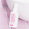 Eine Flasche Love Touch Shine Spray steht auf rosa Fliesen neben einem weißen Waschbecken.