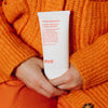 Person in einem orangefarbenen Pullover hält eine weiße Tube Mane Attention Protein Treatment.
