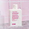 Eine weiße Flasche Mane Tamer Smoothing Shampoo mit Schaum steht auf einer nassen Oberfläche vor einem rosa Fliesenhintergrund.
