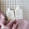 Durch eine beschlagene Glasduschtür sind das Normal Persons Shampoo und der Conditioner vor einer gefliesten Fläche zu sehen.