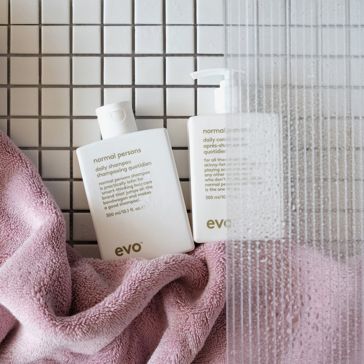 Durch eine beschlagene Glasduschtür sind das Normal Persons Shampoo und der Conditioner vor einer gefliesten Fläche zu sehen.