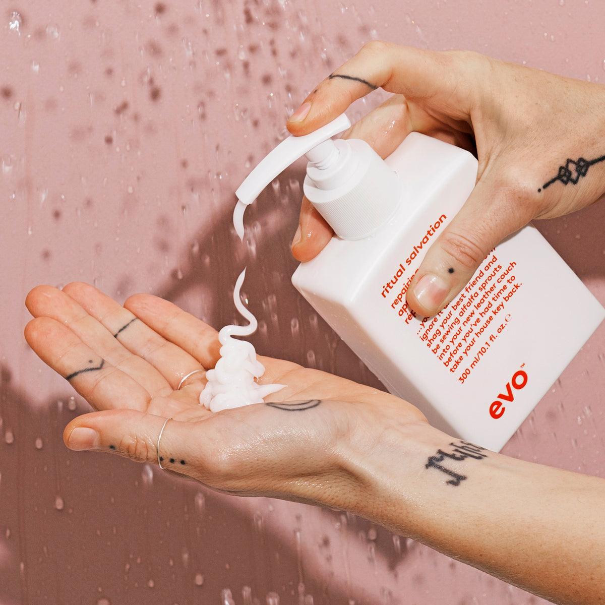 Vor einem nassen rosa Hintergrund pumpt eine Hand weißes Produkt aus einer Flasche Ritual Salvation Repairing Conditioner auf die Andere.