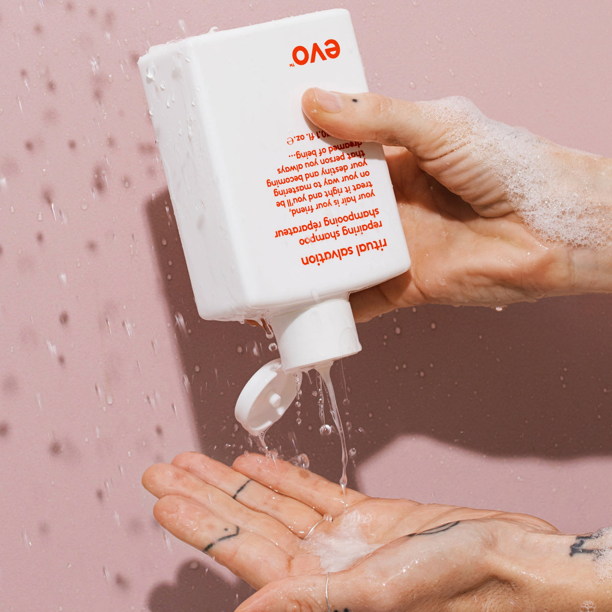 Eine Hand gießt vor rosa Hintergrund Ritual Salvation Repairing Shampoo aus einer weißen Flasche auf ihre Handfläche.