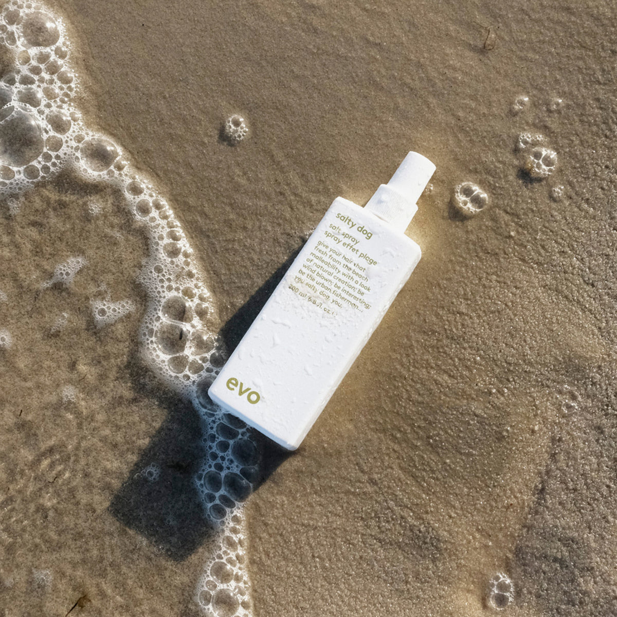 Eine weiße Flasche Salty Dog Salt Spray von Evo liegt auf dem nassen Sandstrand in der Nähe schäumender Wellen.