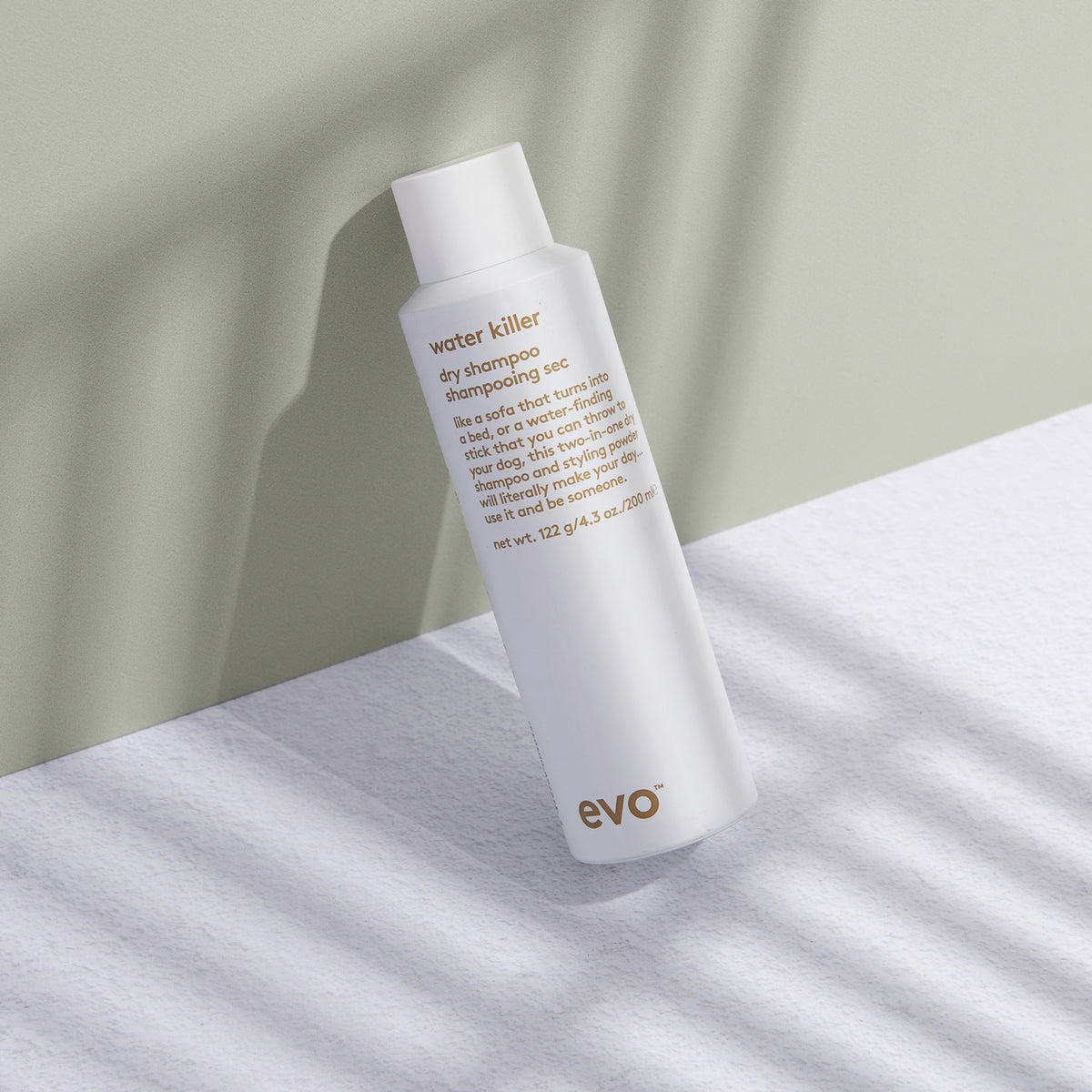 Eine Flasche Water Killer Trockenshampoo wirft einen Schatten auf eine leicht strukturierte Oberfläche und eine grüne Wand.