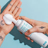 Eine Person verteilt weißen Schaum des Whip It Good Moisture Mousse auf ihrer Handfläche vor blauem Hintergrund.