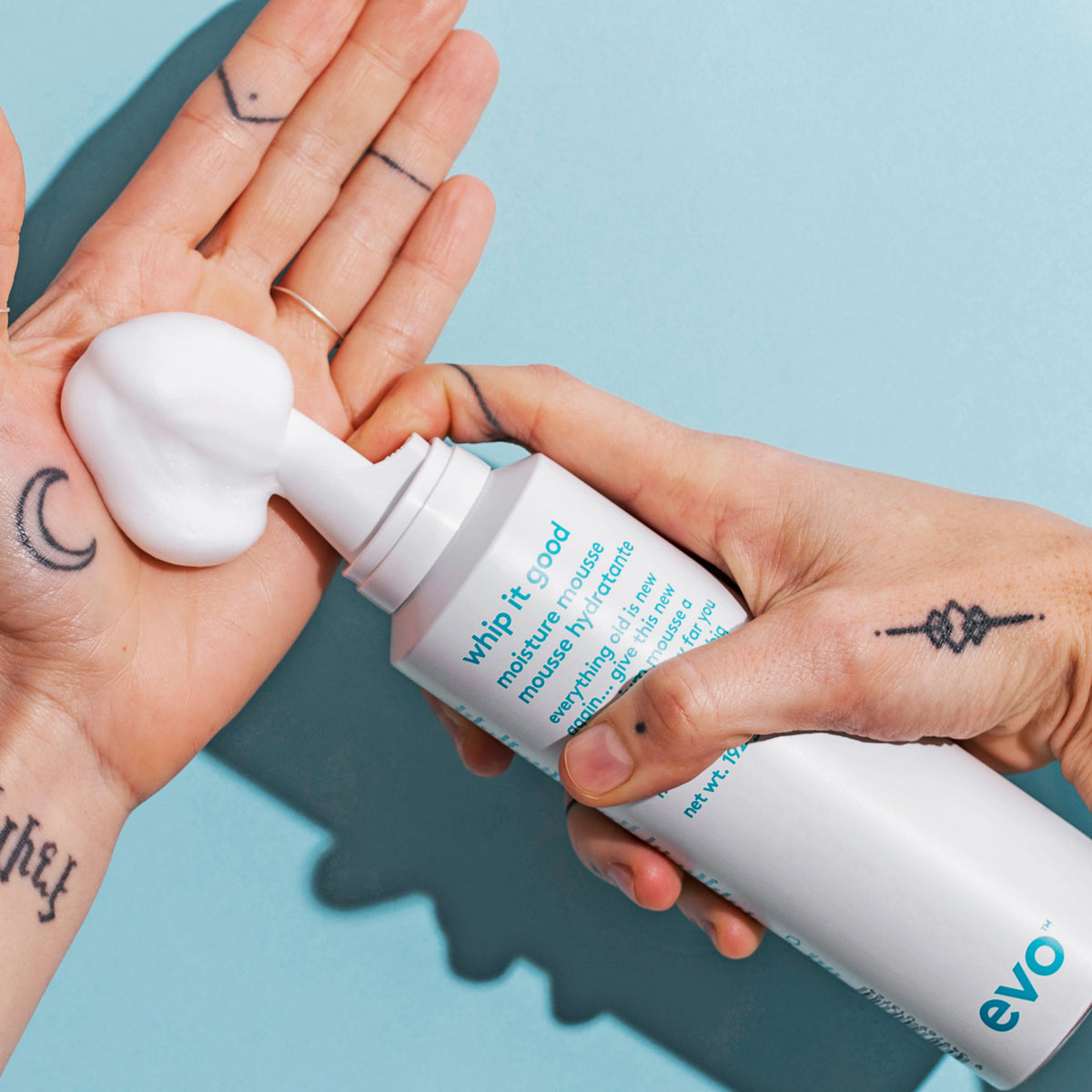 Eine Person verteilt weißen Schaum des Whip It Good Moisture Mousse auf ihrer Handfläche vor blauem Hintergrund.