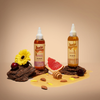 Zwei Flaschen Hair Syrup mit Grapefruit, Honig, Nüssen, Kirschen und einer gelben Blume auf brauner Rinde.