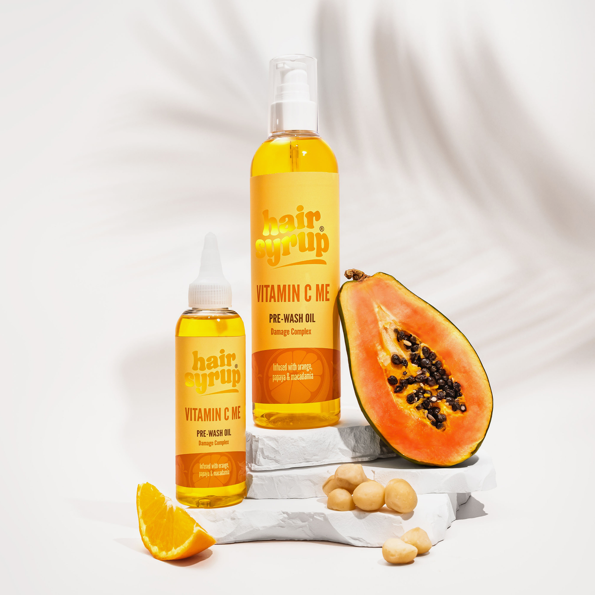 Zwei Flaschen Hair Syrup Vitamin C Me werden mit Papaya, Orangenscheiben und Macadamianüssen auf weißen Steinen präsentiert.