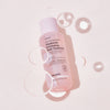Eine rosa Flasche Real Complexion Hyaluron Exfoliating AHA Treatment mit Blasen auf hellrosa Hintergrund.