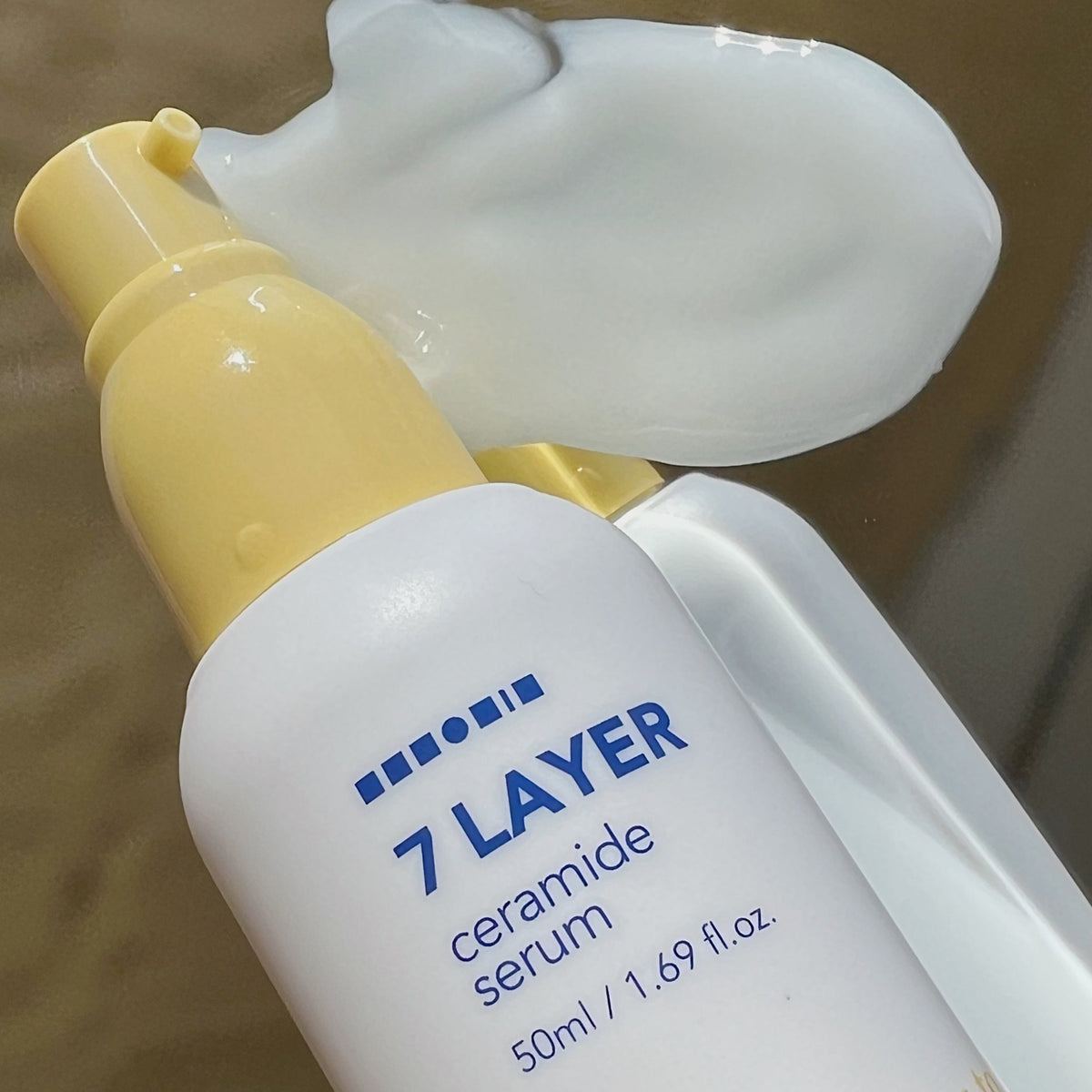 Eine Flasche Seven Layer Ceramide Serum liegt auf der Seite, dahinter ist weißes Serum verteilt.
