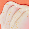 Nahaufnahme der Vitamin C Glow Capsule Lotion ist eine cremeweiße Lotion mit kleinen orangefarbenen Kapslen auf einer korallenfarbenen Oberfläche.