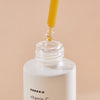 Eine Pipette mit gelbem Serum schwebt über einer weißen Flasche Vitamin C Glow Serum vor einem beigen Hintergrund.