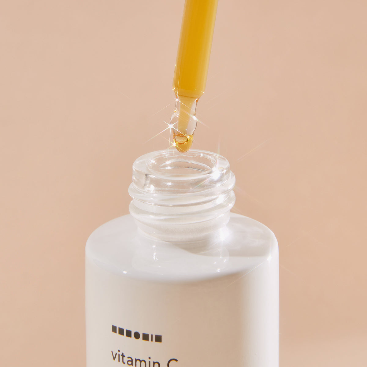 Eine Pipette mit gelbem Serum schwebt über einer weißen Flasche Vitamin C Glow Serum vor einem beigen Hintergrund.