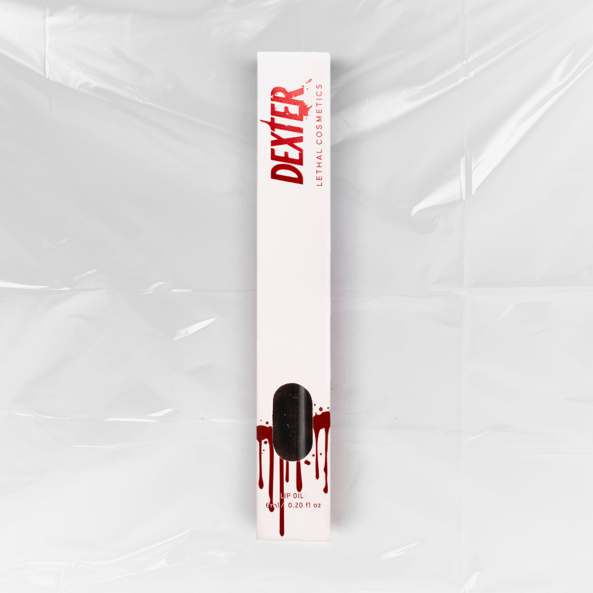 Weiße Schachtel Dexter Lip Oil Aconite von Lethal Cosmetics mit Bluttropfen-Design und Logo auf einem Folienhintergrund.