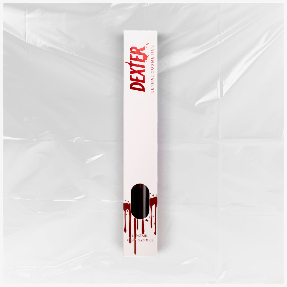 Weiße Verpackung des Dexter Lip Stain Dark Passenger von Lethal Cosmetics mit rotem Text und Bluttropfen-Design vor einer  Plastikfolie.