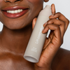 Eine lächelnde Person hält eine Flasche On Set – Long-Lasting Setting Spray von LH Cosmetics an ihr Gesicht.