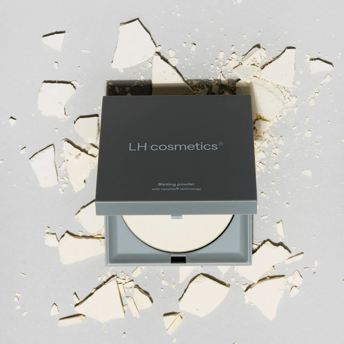 Graue eckige Kompaktdose des Blotting Powder von LH Cosmetics liegt auf verstreutem, zerbrochenem Puder vor einem hellgrauen Hintergrund.
