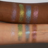Zwei Arme mit unterschiedlichen Hauttönen zeigen vier schimmernde Farbtöne – Lila, Rosa, Blau und Grüngold – aus der Siren Song Palette von LH Cosmetics.