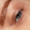 Nahaufnahme eines geschminkten Auges mit blau-grüner Iris und schimmerndem Pfirsichgold-Lidschatten auf Lid, Braue und unterem Augenbereich.