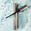 Graue Mascara mit geöffnetem Deckel und sichtbarer Bürste liegt auf einer Wasseroberfläche mit Lichtreflexen; Wassertropfen bedecken die Verpackung.