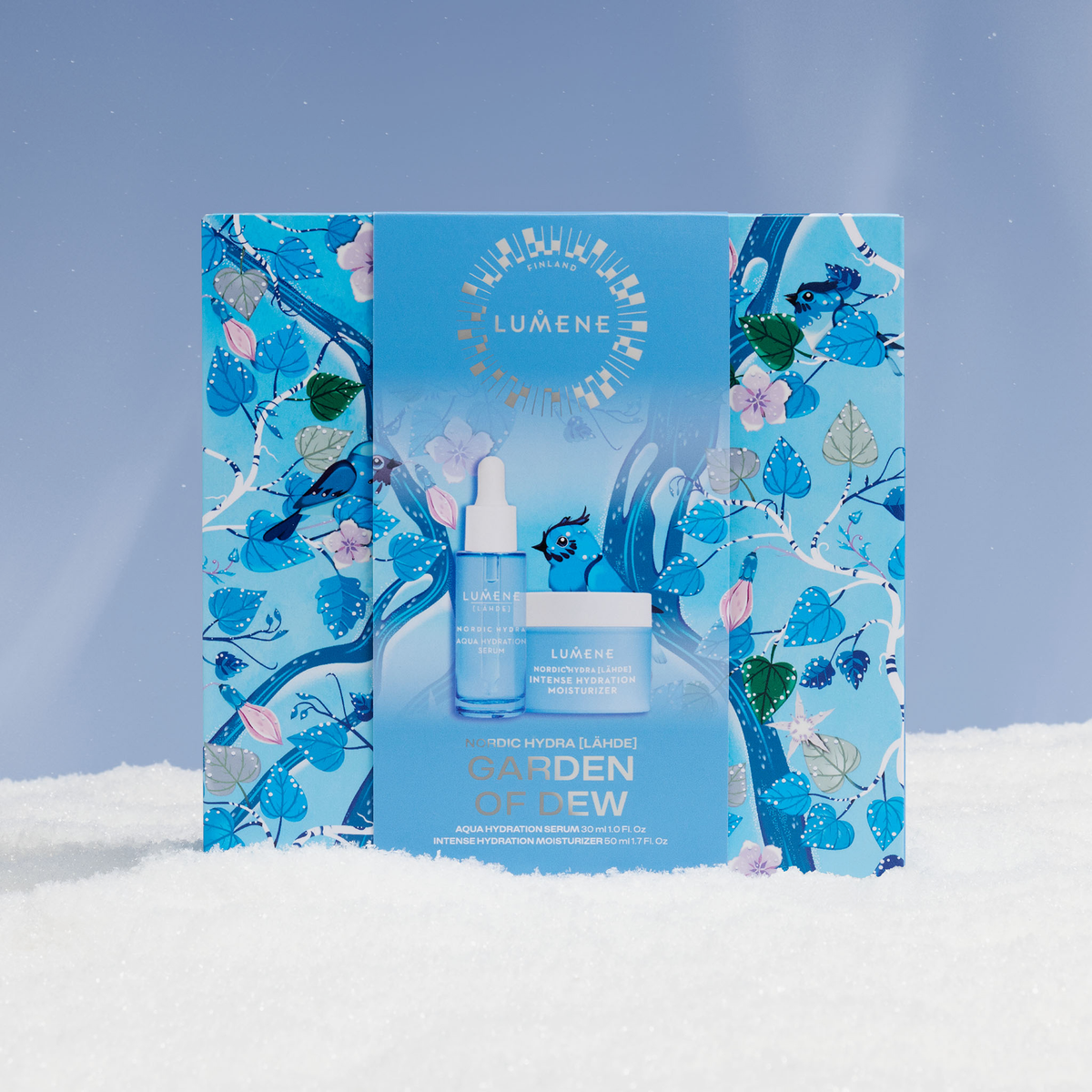 Ein blaues Lumene GARDEN OF DEW SET 2026 steht auf Schnee, im Hintergrund sind Blumen- und Vogelmotive zu sehen.
