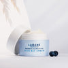 Ein geöffneter hellblauer Tiegel Lumene NORDIC SENSITIVE [HERKKÄ] Rich Day Cream steht auf einer weißen Fläche, daneben zwei Blaubeeren.