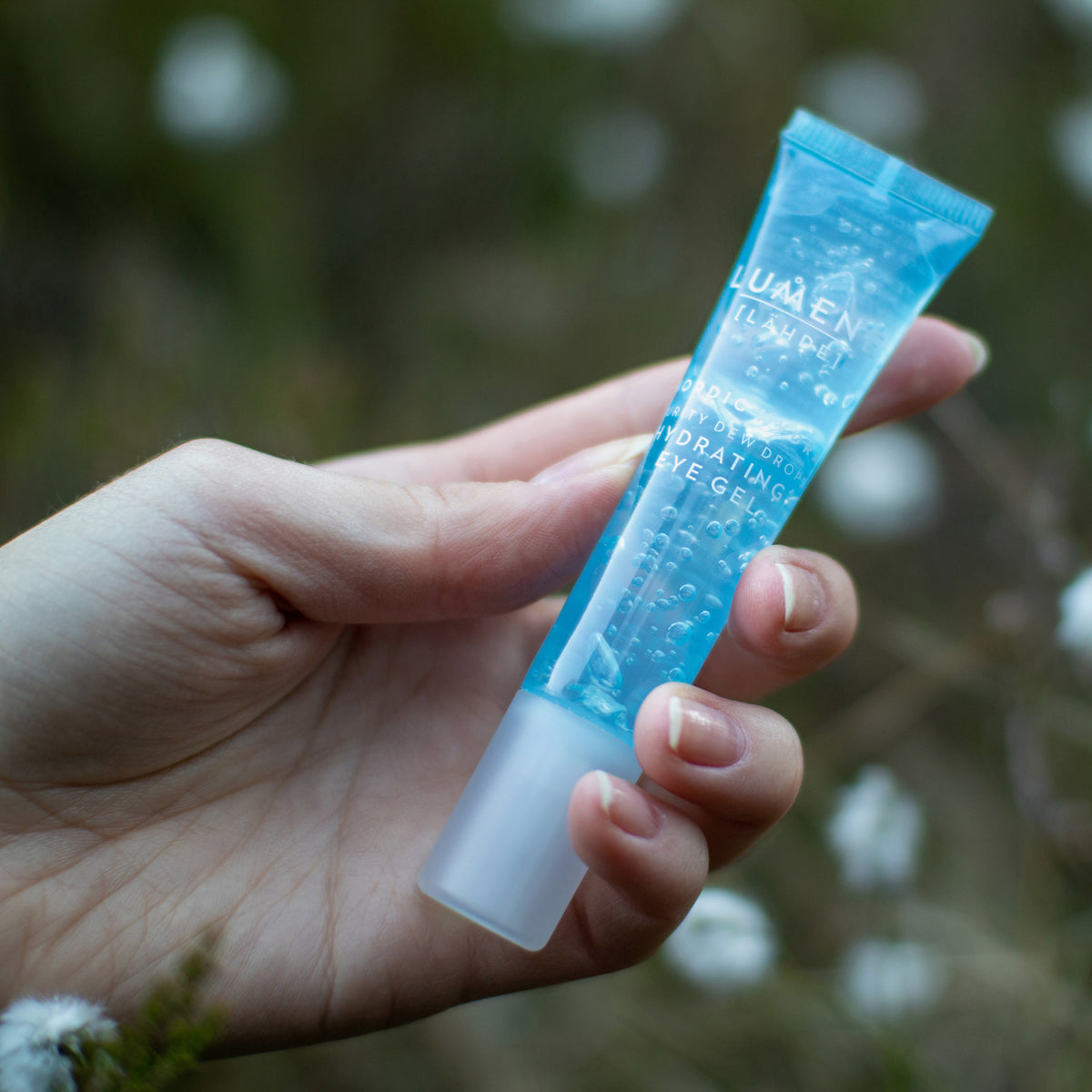 Eine Hand hält eine kleine blaue Tube Lumene NORDIC HYDRA [LAHDE] Purity Dew Drops Hydrating Eye Gel im Freien, vor einem verschwommenen grünen Hintergrund.