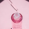 Ein Tropfer gibt Lumene NORDIC BLOOM [LUMO] Vegan Collagen Essence in die Flasche vor einem zartrosa Hintergrund.