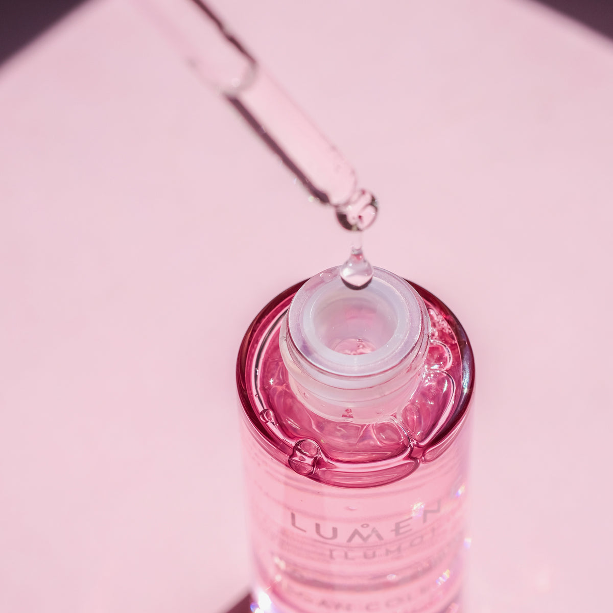 Ein Tropfer gibt Lumene NORDIC BLOOM [LUMO] Vegan Collagen Essence in die Flasche vor einem zartrosa Hintergrund.