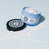 Geöffneter blauer Tiegel der maria nila Coils & Curls Finishing Treatment Masque mit weißer Creme, die auf einer hellblau gefliesten Fläche verteilt ist.