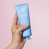 Eine Hand hält eine blaue Tube des maria nila Coils & Curls Luscious Curl Definer vor einem hellrosa Hintergrund.